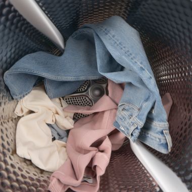 Blick in die Trommel einer Waschmaschine. In der Trommel liegen verschiedene Kleidungsstücke, eine blaue Jeans, ein helles und ein rosafarbenes Textil, locker übereinander. Die Trommelstruktur und der Mitnehmer sind deutlich sichtbar.