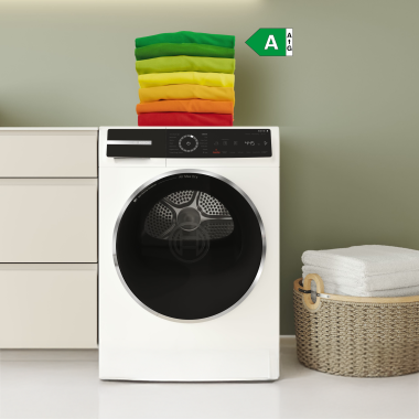 Frontansicht einer weißen Waschmaschine. Darauf liegen farbig sortierte Handtücher von grün bis rot. Rechts daneben steht ein geflochtener Wäschekorb mit weißen Handtüchern, daneben ein grünes Energielabel mit der Kennzeichnung „A“.