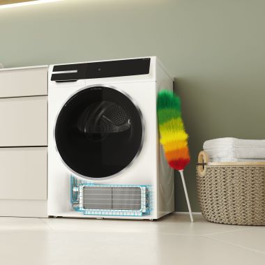 Weiße Waschmaschine in einem hellen Raum. Unten an der Maschine ist ein herausgezogener Kondensatorfilter zu sehen. Rechts ein bunter Staubwedel, daneben steht ein geflochtener Wäschekorb mit gefalteten weißen Handtüchern.