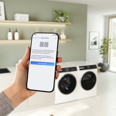Eine Hand hält ein Smartphone. Auf dem Display ist eine Programm-App zu sehen. Im Hintergrund stehen unscharf eine Waschmaschine und ein Trockner in einem hellen, modern eingerichteten Raum mit Regalen, Pflanzen und Pflegeprodukten.