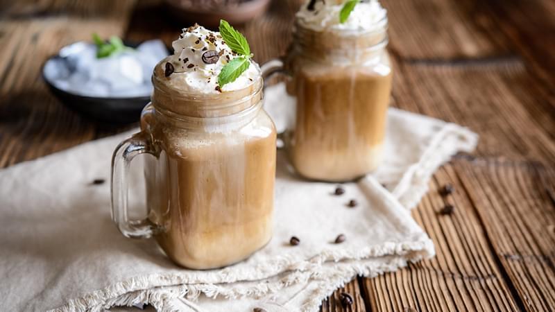 Zwei Gläser mit Kaffee-Frappé, dekoriert mit einer großzügigen Portion Sahne stehen auf einem Holztisch