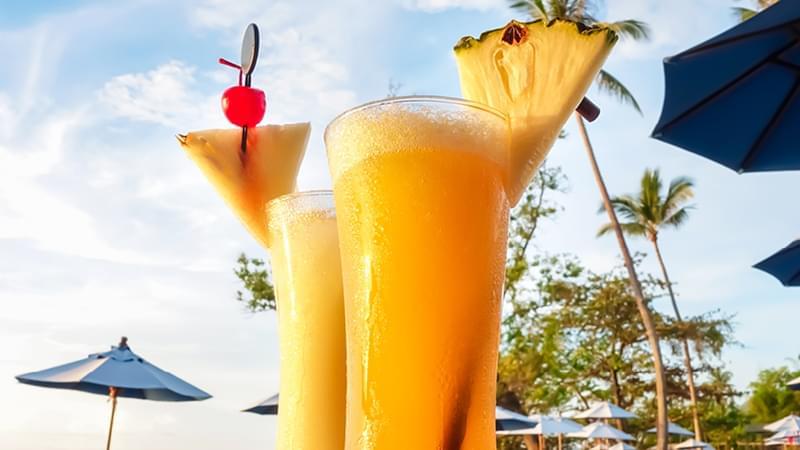 Zwei Gläser eines tropischen Slush Getränks mit Ananas auf einem Tisch am Strand, umgeben von einer malerischen Küstenlandschaft.