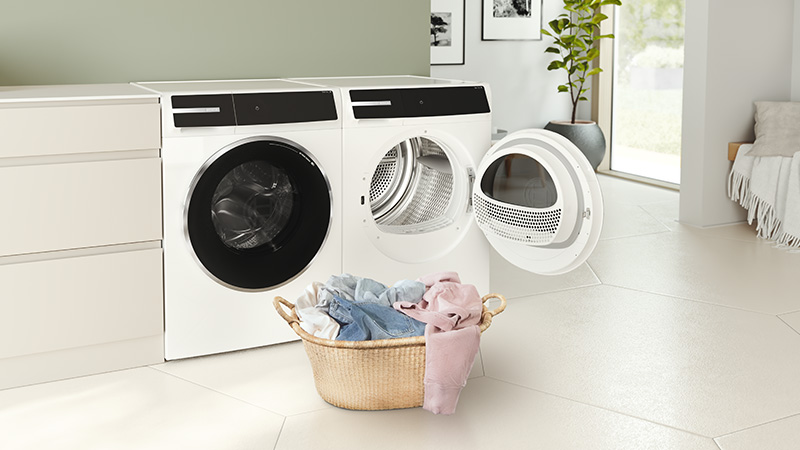Bosch Waschmaschine und geöffneter Trockner in einem hellen Hauswirtschaftsraum. Vor den Geräten steht ein Wäschekorb mit frisch gewaschener Kleidung, der Trockner ist zum Beladen geöffnet.